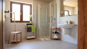A modern bathroom with a glass shower enclosure, a wooden chair, a small shelf with toiletries, and a window with blinds.