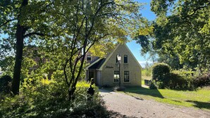 Ein traditionelles Haus mit einem geneigten Dach, einer Ziegelaußenwand und einem sichtbaren Schornstein. Eine gepflasterte Auffahrt führt zum Eingang, umgeben von üppigem Grün und Bäumen.