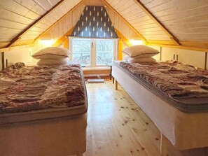 A small attic bedroom with two single beds, a window with a leaf-patterned valance, and wooden walls.