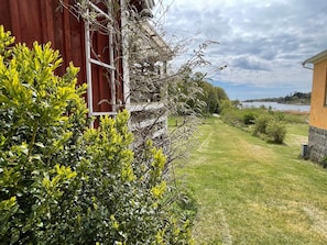 A red building with a white window, surrounded by green shrubs and a grassy area leading to a body of water. - 4 Star Holiday Home in Ronneby (Ronneby)