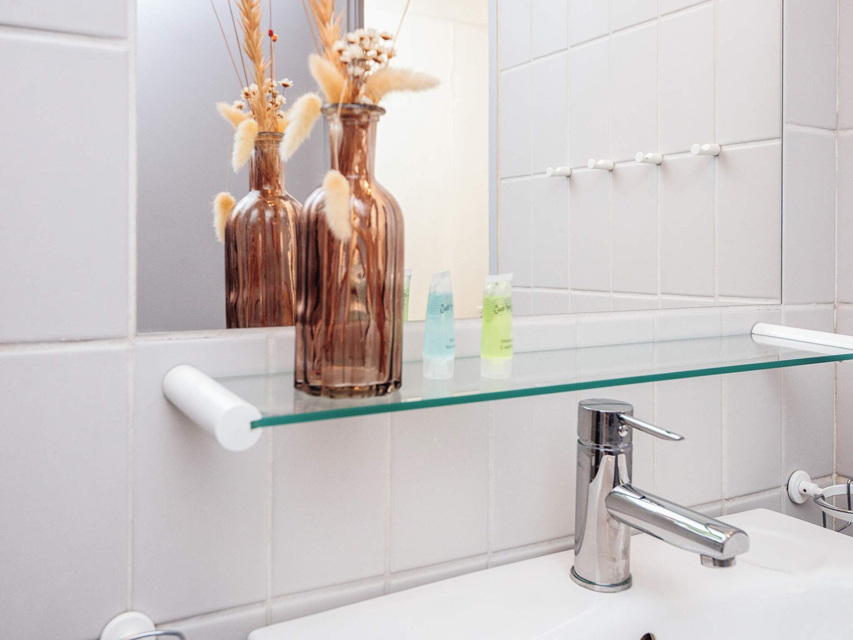 Une salle de bain avec une étagère en verre où sont rangés les articles de toilette, un miroir et un robinet chromé.