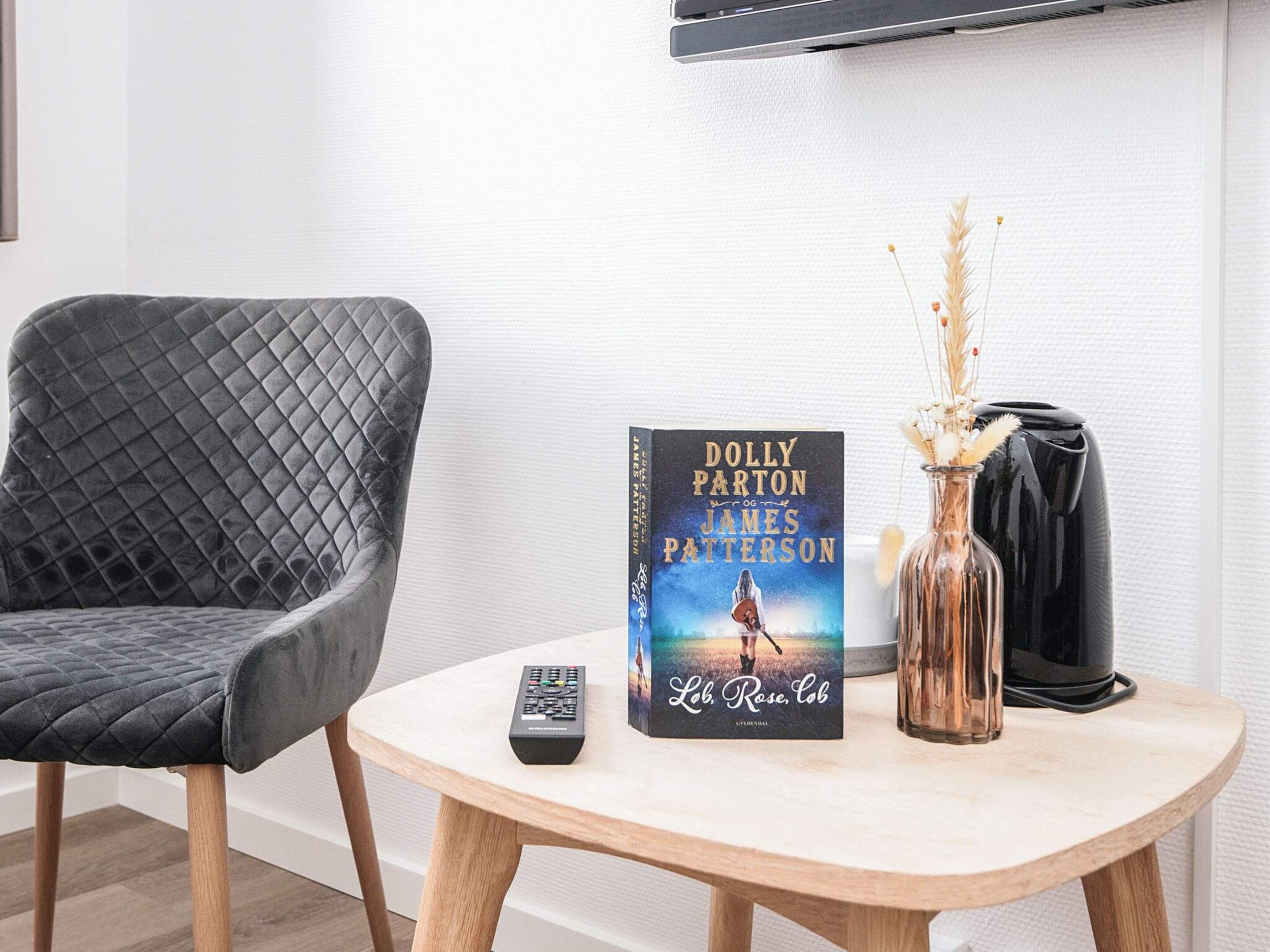 Una habitación con una silla tapizada de gris, una pequeña mesa redonda de madera con un libro titulado 'Lob, Rose, Cob' de Dolly Parton y James Patterson, un control remoto y un florero con plantas secas.