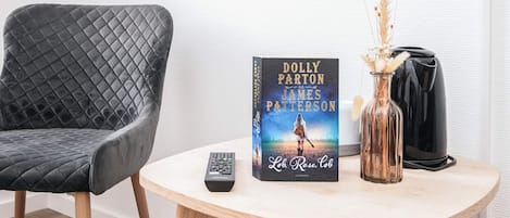 Una stanza con una poltrona grigia imbottita, un piccolo tavolino rotondo in legno con un libro intitolato 'Lob, Rose, Cob' di Dolly Parton e James Patterson, un telecomando e un vaso con piante essiccate.
