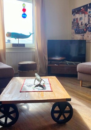 Un soggiorno con un tavolino in legno, una televisione su un mobile e una finestra con un decoro appeso.