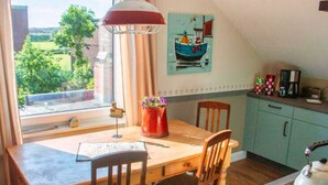 A dining area with a wooden table, chairs, a red vase with flowers, and a view of the outdoors through a window. - Bunte Kuh Comfortable Holiday Residence (Borkum)