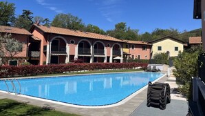 A swimming pool with a clear blue water surface, surrounded by a well-maintained lawn and a row of buildings with arched balconies. - Perlago Comfortable Holiday Residence (Costermano sul Garda)