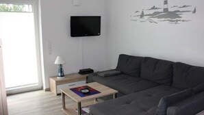 A modern living room with a grey sofa, a flat-screen TV mounted on the wall, a small wooden table with a lamp, and a decorative wall art of a lighthouse. - Holiday Apartment Cabin 2c (Norden)