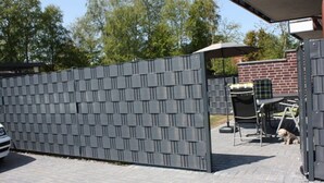 A gated outdoor area with a paved patio, a barbecue setup, and a brick building in the background. - Holiday Apartment Cabin 2c (Norden)
