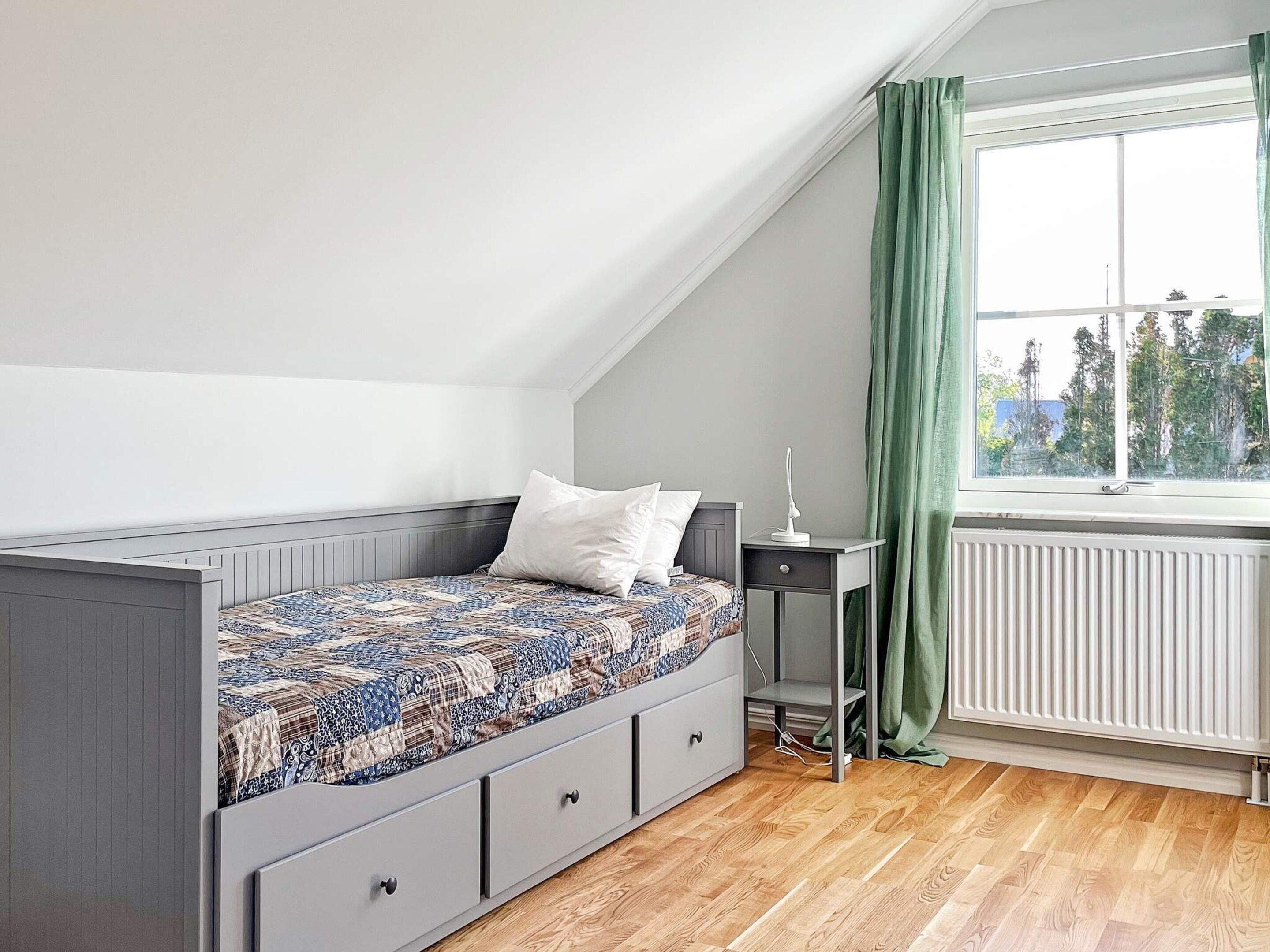 A bedroom with a sloped ceiling, a bed with patterned bedding, a bedside table with a lamp, and a window with curtains.