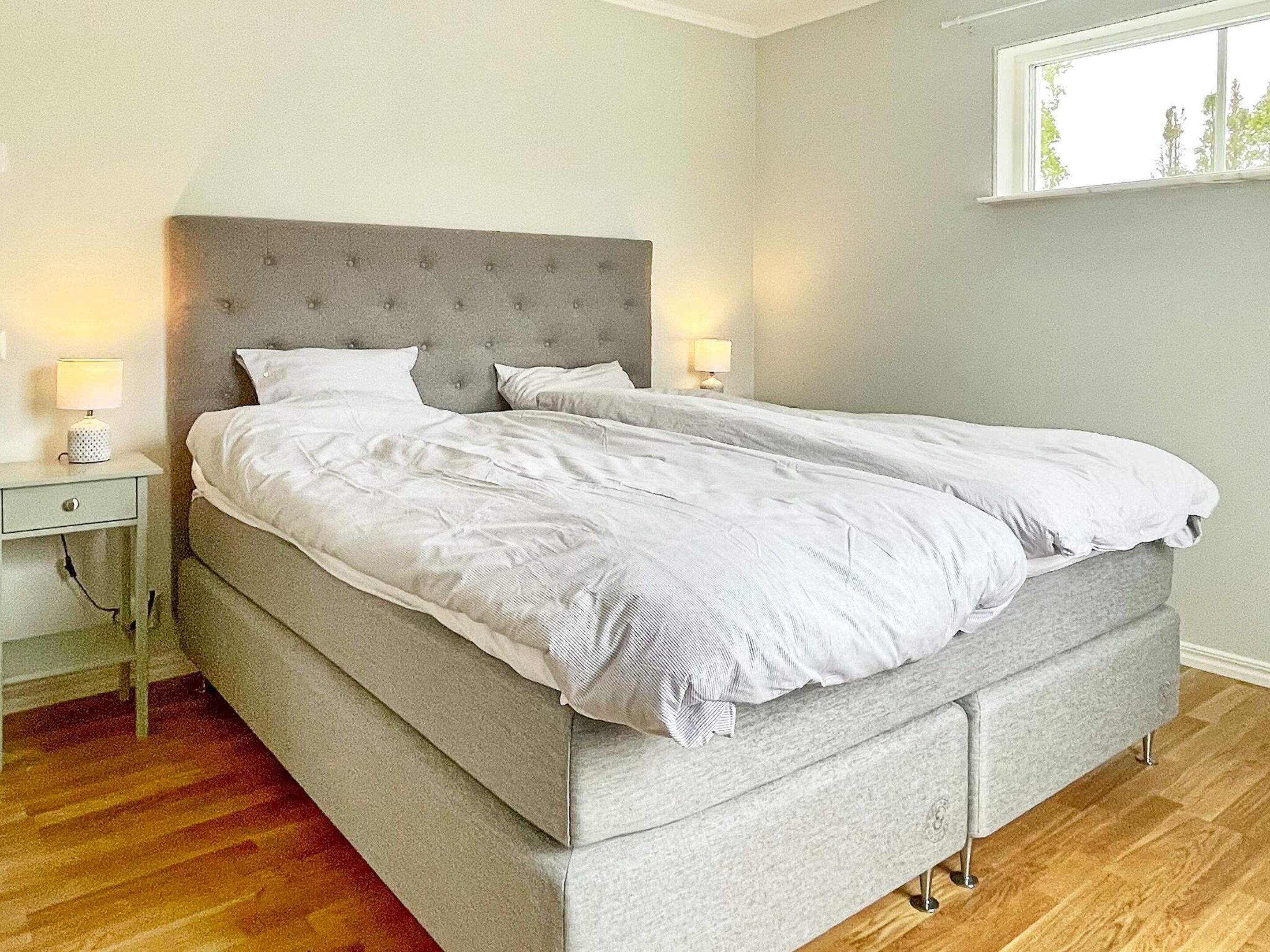 A neatly made bed with a grey headboard, two bedside lamps, and a wooden floor.