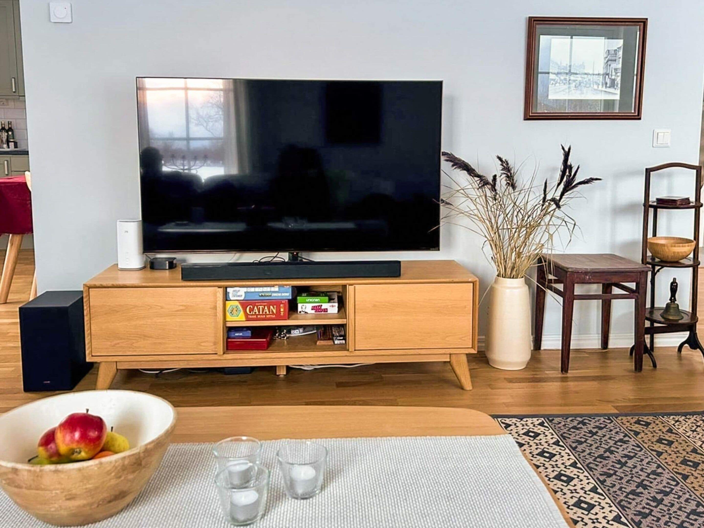 Una sala de estar con un televisor de pantalla plana sobre un soporte de madera, una estantería con juegos de mesa, una mesa de centro de madera con un cuenco de fruta y vasos, y una alfombra estampada.