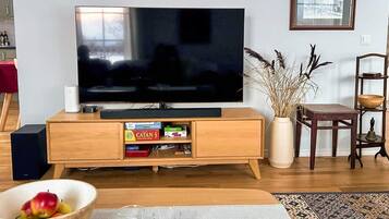 Una sala de estar con un televisor de pantalla plana sobre un soporte de madera, una estantería con juegos de mesa, una mesa de centro de madera con un cuenco de fruta y vasos, y una alfombra estampada.