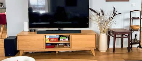 A living room with a flat-screen TV on a wooden stand, a bookshelf with board games, a wooden coffee table with a bowl of fruit and glasses, and a patterned rug.