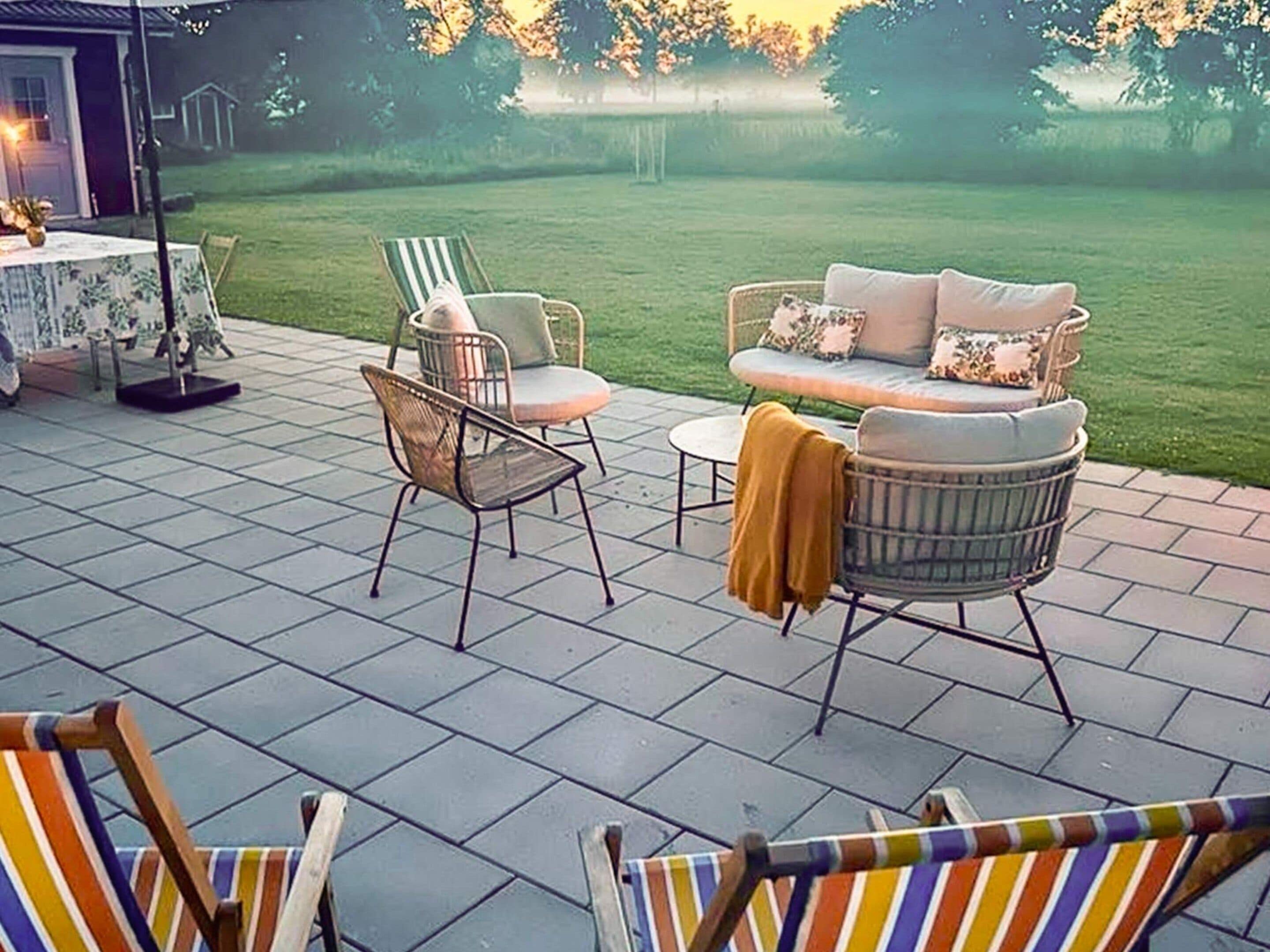 An outdoor patio with a sofa and chairs, a table with a vase, and a view of a grassy field.