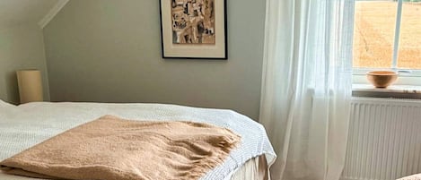 A bedroom with a bed, a framed picture on the wall, a window with a view of a field, and a radiator.