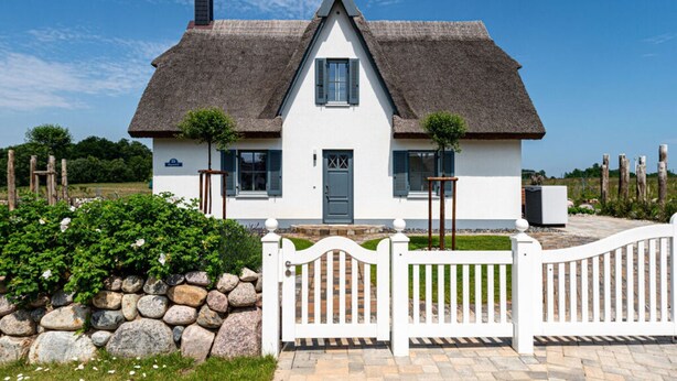 Een wit vakantiehuisje met rieten dak, een stenen muur en een witte picket fence.