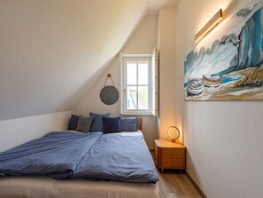 A bedroom with a bed, a bedside table with a lamp, a window, and a painting on the wall.