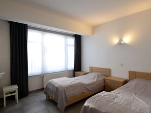 A hotel room with two beds, a small bedside table, a chair, and a window with curtains.