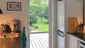 Ett kök med en diskho i rostfritt stål, en kaffebryggare, ett kylskåp och en träveranda som syns genom dörren.
