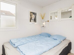 A single bed with a blue blanket and pillows, a bedside table with a lamp, a framed picture on the wall, and a window with a view of a building. - 6 Person Holiday Home in Vestervig (Vestervig)