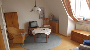 A cozy room with a wooden floor, a round table with a white tablecloth, a wicker chair, a wooden cabinet, a television on a wooden stand, and a window with curtains. - Lagoon on the Wadden See on Wangerooge (Wangerooge)