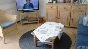 A living room with a television displaying a news channel, a wicker chair, a wooden table with a white tablecloth, a blue sofa, and a wooden cabinet with books and decorative items. - Lagoon on the Wadden See on Wangerooge (Wangerooge)