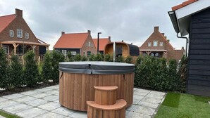 A wooden hot tub with steps, surrounded by a paved patio and lush greenery.