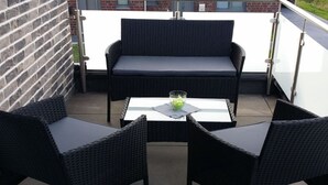 A balcony with a wicker sofa and two chairs, a glass table with a vase, and a brick wall in the background.