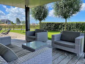 Un patio al aire libre con mobiliario de mimbre, una mesa con superficie de vidrio y vistas a un paisaje verde con seto y árboles.