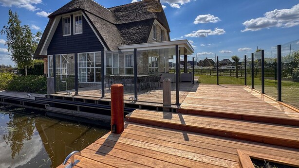 A house with a large deck, a glass conservatory, and a view of a canal. - Villa by Tjeukemeer With Sauna & Whirlpool (Delfstrahuizen)