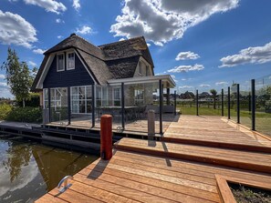 A house with a large deck, a glass conservatory, and a view of a canal. - Villa by Tjeukemeer With Sauna & Whirlpool (Delfstrahuizen)