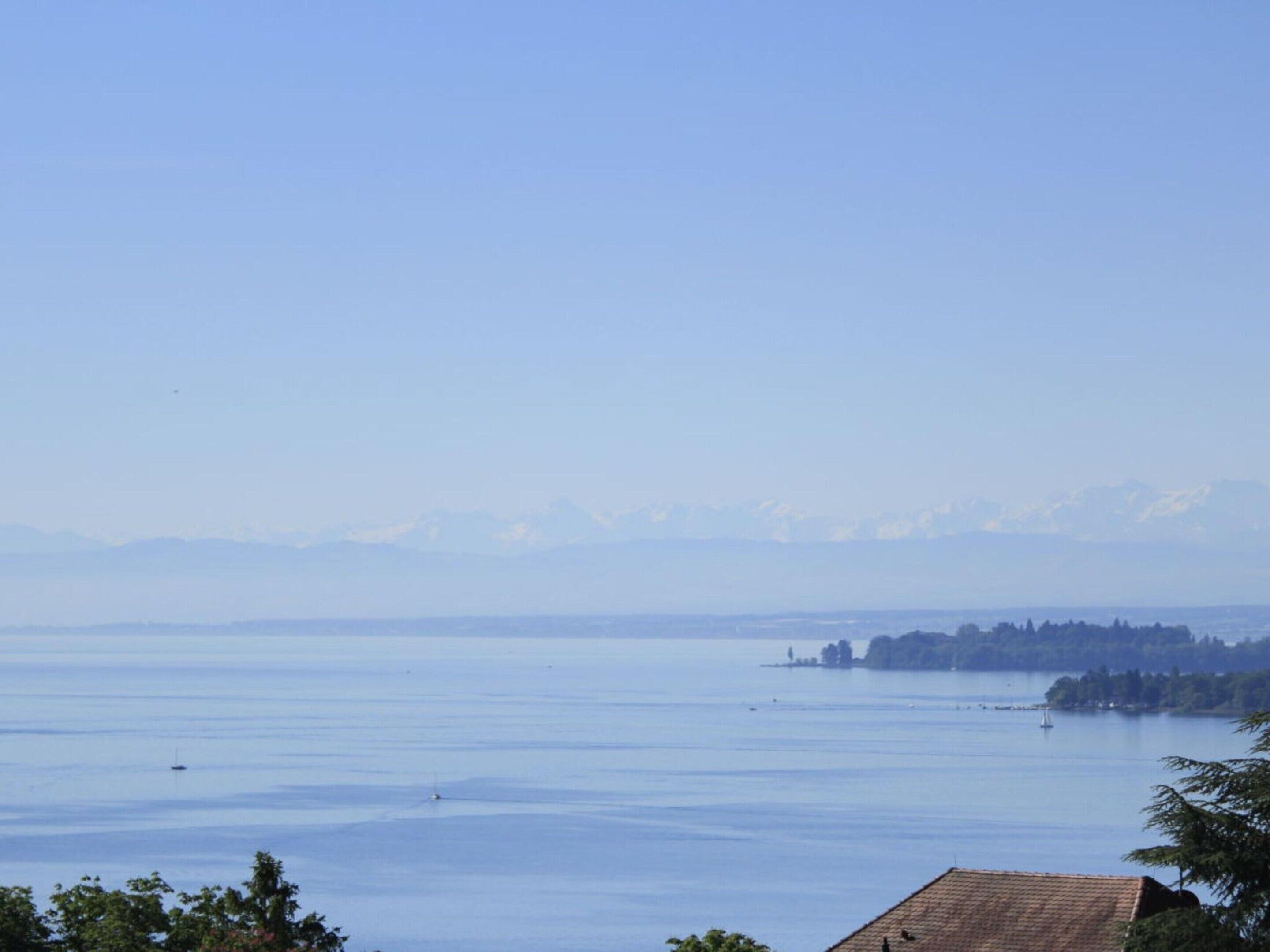 A serene lake with a distant mountain range, a small island with trees, and a building with a tiled roof in the foreground.