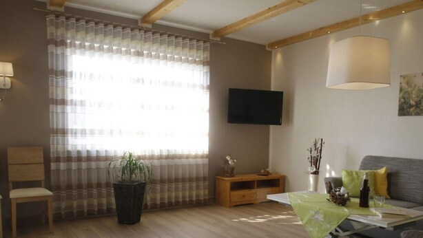 A modern living room with a wooden floor, a flat-screen TV mounted on the wall, a sofa, a small wooden side table, and a potted plant. - Tasteful Apartment in Burg (Burg (Spreewald))