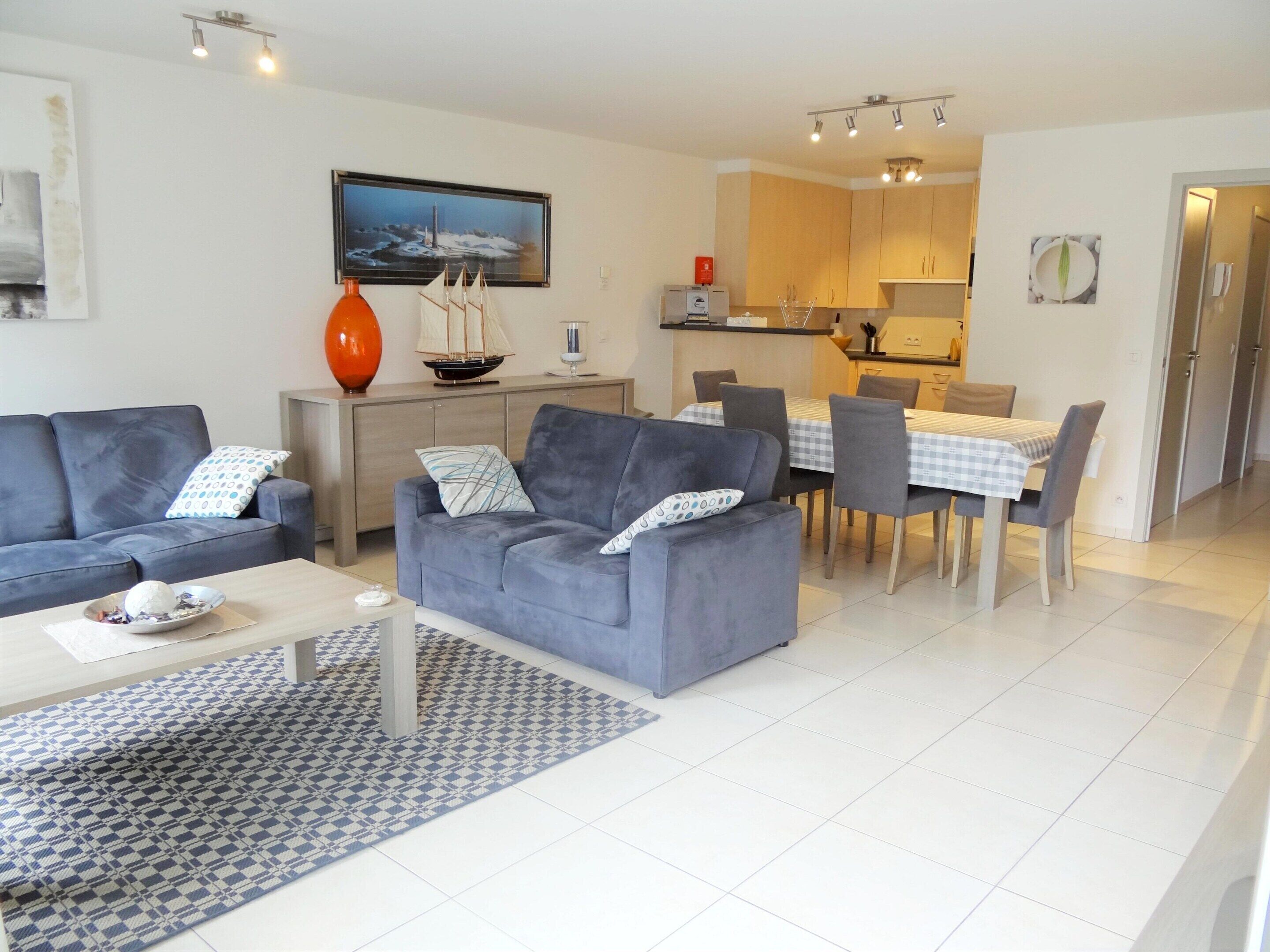 A modern living room with a grey sofa, a coffee table, and a dining area with a checkered tablecloth.
