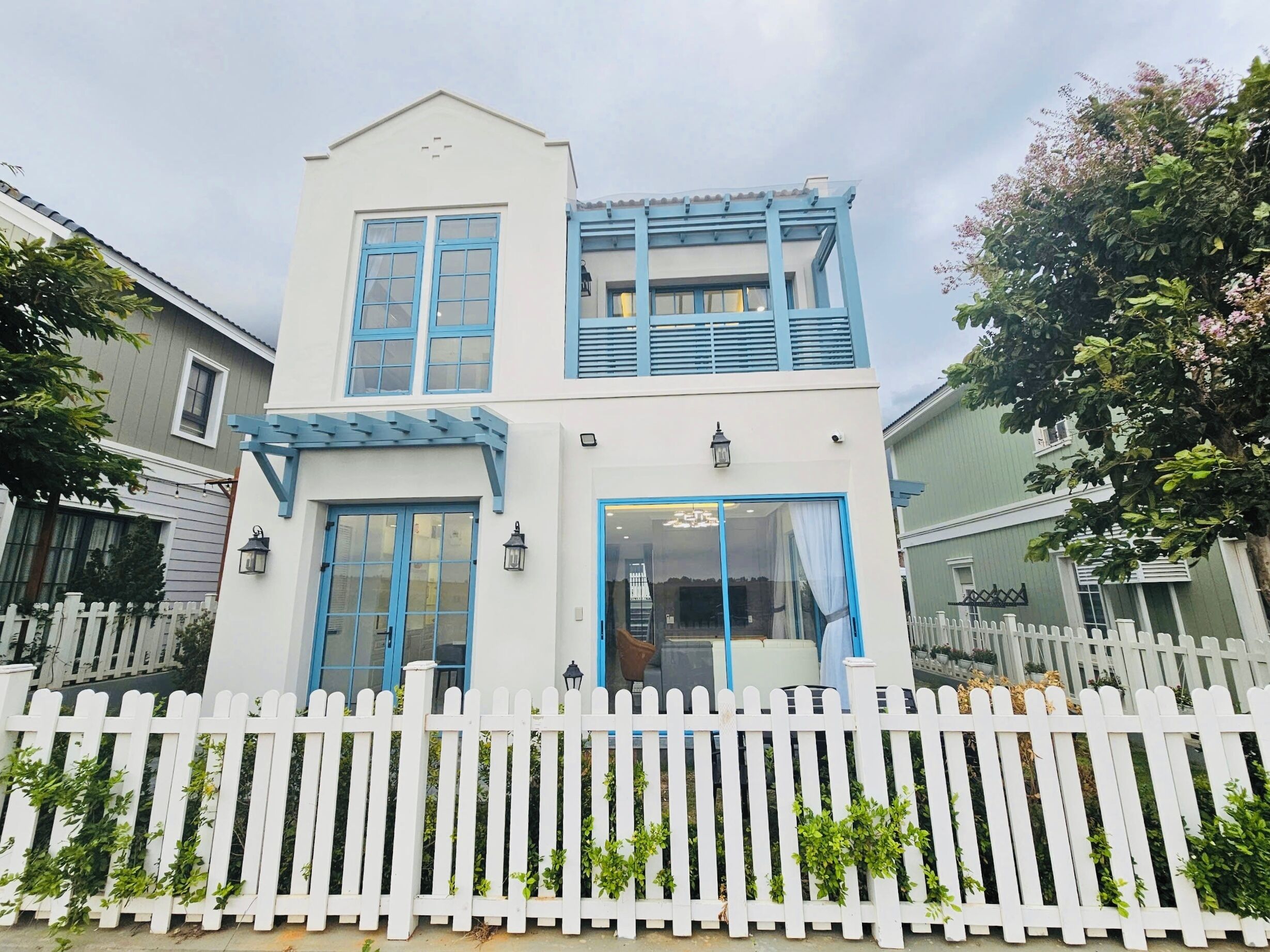 Una casa de dos pisos con fachada blanca y detalles azules, una valla de piquetes blancos y un jardín con plantas en macetas.