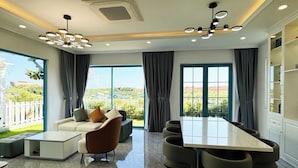 A modern dining area with a large table and chairs, a sofa, and a view of a garden through large windows. - Holiday Villa Novaworld Phan Thiet (Tien Thanh)