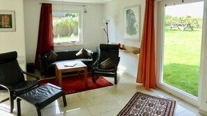 A living room with a sofa, chairs, a coffee table, and a view of a garden through a sliding door. - Holiday Apartment Sandpiper Ground Floor (Klütz)