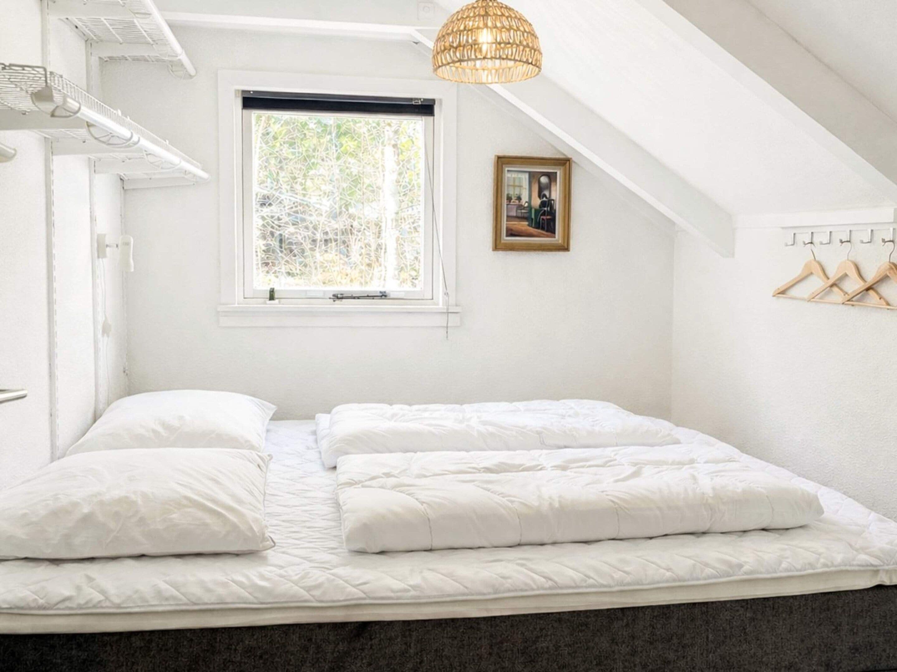 Ein Schlafzimmer mit Bett, Fenster, Pendelleuchte und Kleiderhaken.