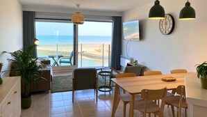 A modern living room with a large window overlooking a beach, a dining area with wooden furniture, and a flat-screen TV mounted on the wall. - Apartment in Nieuwpoort With Sea Views (Nieuwpoort)