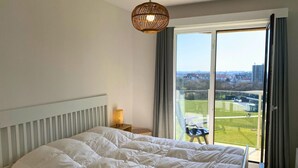 A bedroom with a large bed, a hanging light fixture, a nightstand, and a view of a green landscape through a sliding glass door. - Apartment in Nieuwpoort With Sea Views (Nieuwpoort)