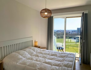 Une chambre dotée d’un grand lit, d’un luminaire suspendu, d’une table de chevet et offrant une vue sur un paysage verdoyant grâce à une porte-fenêtre coulissante.
