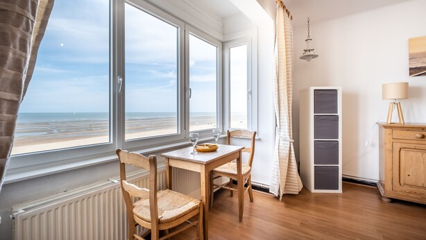 A room with a wooden table and chairs, a radiator, a cabinet, and a view of the beach through large windows. - Apartment on Belgian Coast With Sea View (Koksijde)