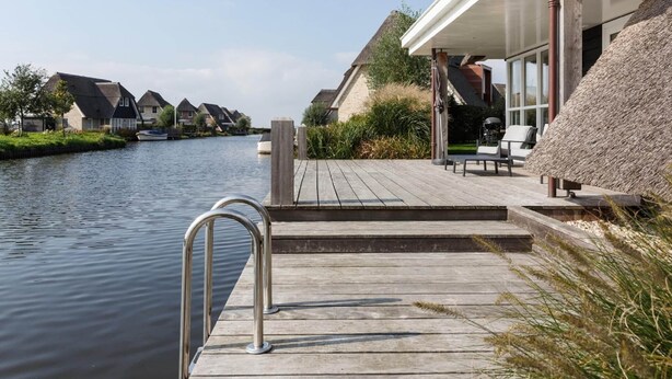 A wooden deck with a metal railing overlooking a calm river, surrounded by traditional houses with thatched roofs.