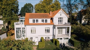 Een groot, wit herenhuis met een rode pannendak, veel ramen en een verzorgde tuin met vormgesnoeide heggen en een pad.
