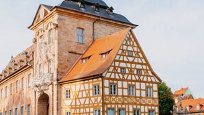 En historisk byggnad med ett markant torn och ett traditionellt hus med korsvirkesfasad, beläget intill en stenbro.