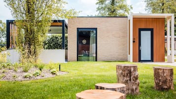 Un edificio moderno con fachada de ladrillo, puerta de vidrio y puerta de madera. Hay árboles y un área verde con troncos de madera.