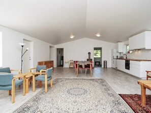 A spacious living area with a dining table, chairs, a rug, and a kitchen with white cabinets and a stove. - 4 Star Holiday Home in Melby (Melby)