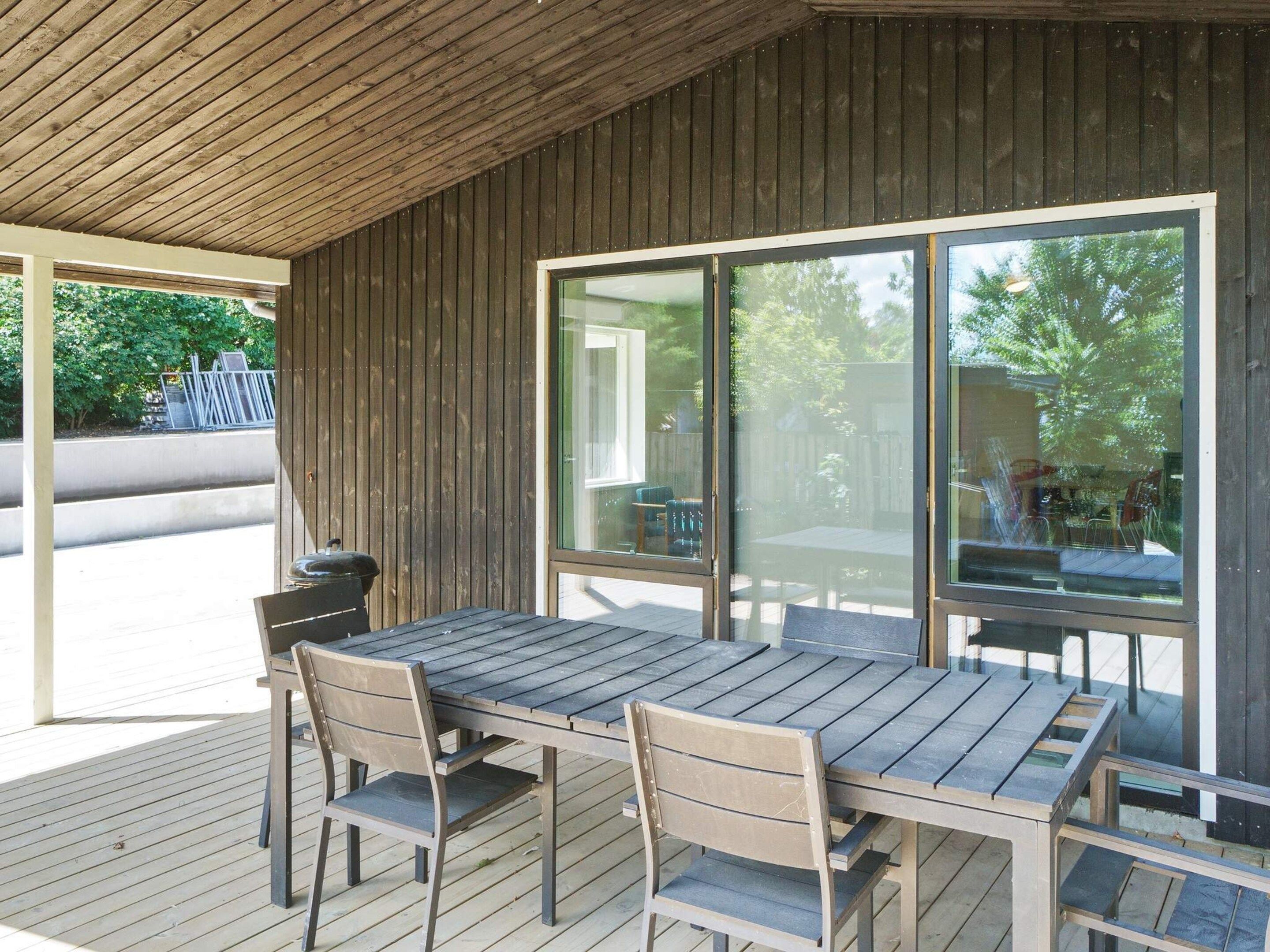 An outdoor patio with a wooden table and chairs, large windows, and a view of the outdoors.