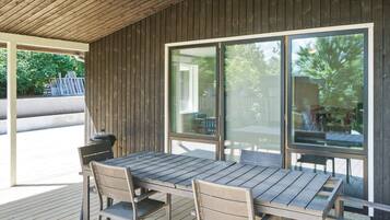 An outdoor patio with a wooden table and chairs, large windows, and a view of the outdoors.