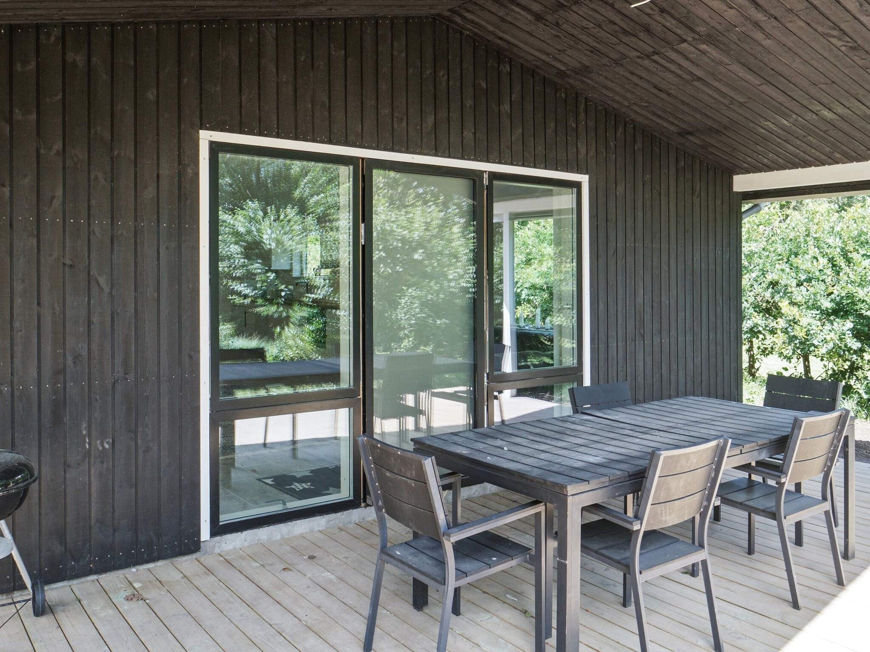 An outdoor patio with a wooden table and chairs, large sliding glass doors, and a view of greenery.